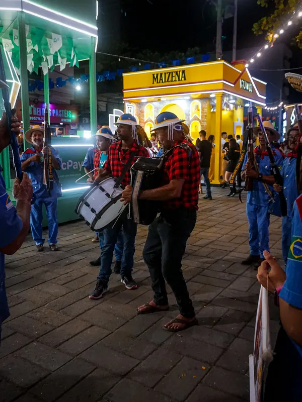 Trio de forró tocando na rua à noite, com zabumba, sanfona e triângulo, de chapéu de couro