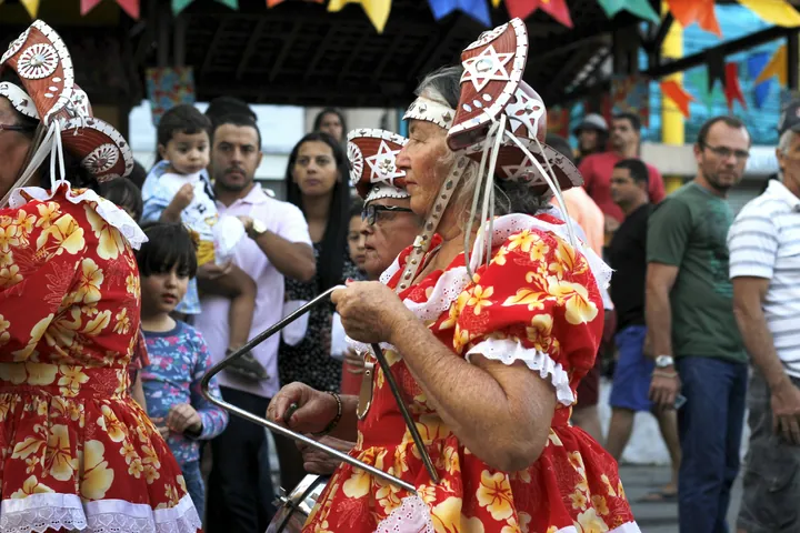 Senhora de vestido de chita vermelho e chapéu de couro tocando triângulo numa festa